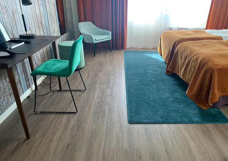 Hotellrum med skrivbord och grön stol, blå matta, två bäddar med rostfärgade filtar samt ljusinsläpp från stort fönster med orange gardiner.